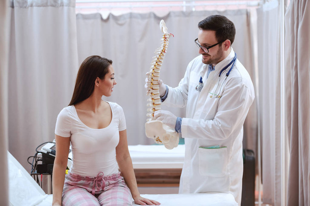 Jovem sorridente branco médico masculino em branco uniforme segurando modelo de coluna vertebral e mostrando a seu paciente
. - Foto, Imagem