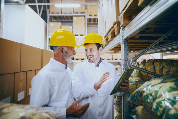 Два улыбающихся кавказских сотрудника в белой форме и желтых шлемах на головах, проверяющих товар на складе. Молодой показывает, что на складе с помощью планшета
. - Фото, изображение