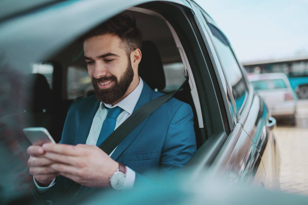 Young smiling handsome Caucasian businessman in blue suit sitting in his expensive with seat belt on and using smart phone. - Fotoğraf, Görsel