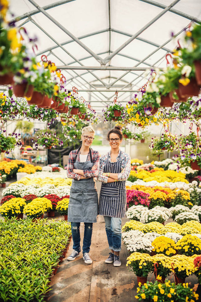 Dos floristas caucásicos en delantales de pie en invernadero con los brazos cruzados rodeados de flores
. - Foto, imagen