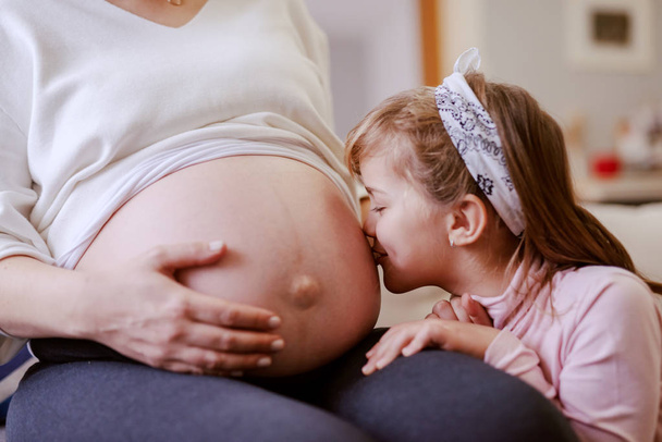 Cute little girl kissing mother's belly. Living room interior. - Photo, Image