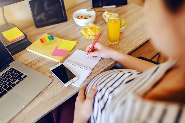Terhes nő ül az asztalnál, és írásban a notebook. Az asztalon egészséges reggeli, laptop és okos telefon. Munka terhes nő koncepció. - Fotó, kép