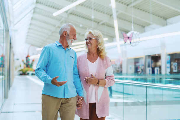 Linda pareja de ancianos tomados de la mano y charlando mientras camina en el centro comercial
. - Foto, Imagen