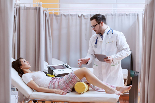 Młody kaukaski lekarz w białym mundurze trzyma tabletkę i wyjaśnia proces regeneracji pacjenta. Pacjent o urazie nóg i leżącej na łóżku. Wnętrze szpitala. - Zdjęcie, obraz