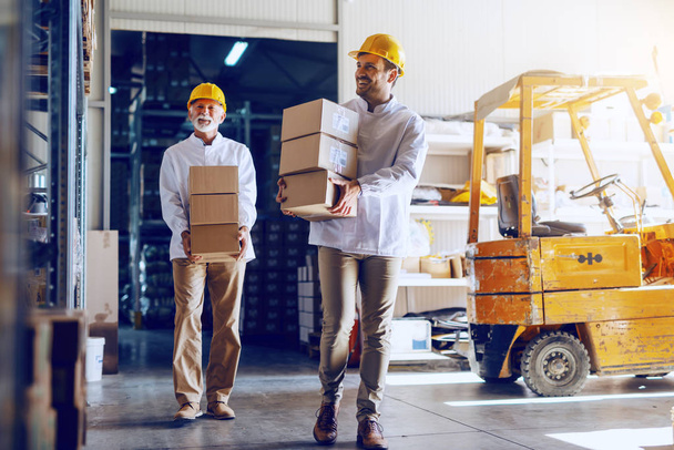 Два рабочих в белых мундирах и в жёлтых шлемах на головах, перемещающих тяжёлые коробки на склад.
. - Фото, изображение