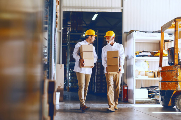 Dos trabajadores de cuello azul en uniformes blancos y con cascos amarillos en cabezas reubicando cajas pesadas en el almacén
. - Foto, Imagen