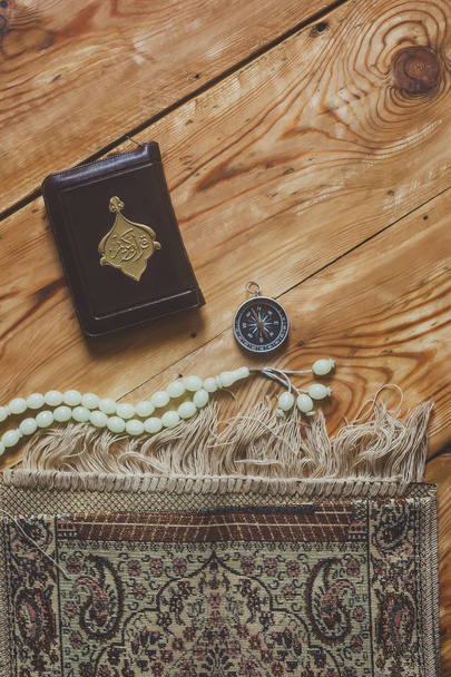 Translation: The Holy Quran. Traditional muslim prayer set bundle. Praying carpet, rosary beads, little version of the Holy Quran and qibla compass on wooden background. Free Space - Photo, Image