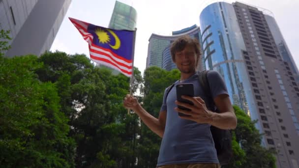 Slowmotion shot of a young man that holds a smartphone and waves Malaysian flag with skyscrapers at a background. Travel to Malaysia concept - Video, Çekim