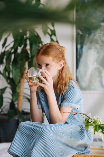 Teenage redhead girl is sitting on a box, drinking tea in a workshop. - Fotoğraf, Görsel