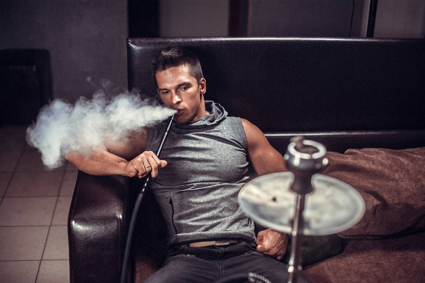 Sexy young man smoking a hookah and smoke issues from the mouth at arabic restaurant. - Photo, Image