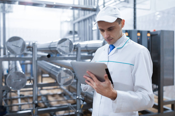 Portrait of man in a white robe and a cap standing in production department of dairy factory with grey tablet - Photo, Image