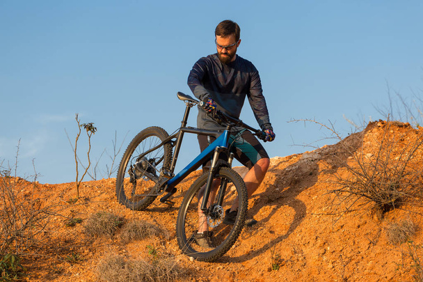 Cyclist in shorts and jersey on a modern carbon hardtail bike with an air suspension fork rides off-road on the orange-red hills at sunset evening in summer - Photo, Image