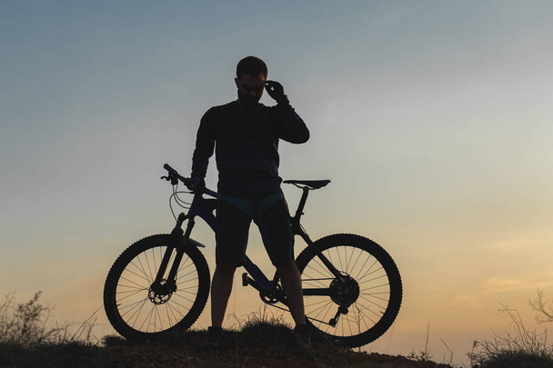 Ciclista en pantalones cortos y jersey en una moderna bicicleta rígida de carbono con una horquilla de suspensión neumática pasea fuera de la carretera en las colinas de color rojo anaranjado al atardecer en verano
 - Foto, Imagen