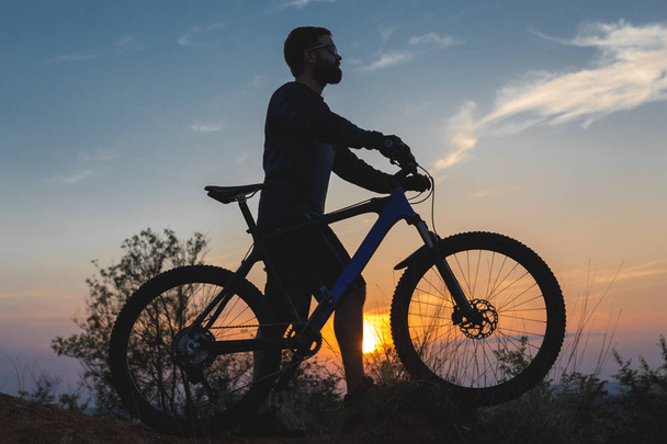 Ciclista en pantalones cortos y jersey en una moderna bicicleta rígida de carbono con una horquilla de suspensión neumática pasea fuera de la carretera en las colinas de color rojo anaranjado al atardecer en verano
 - Foto, imagen