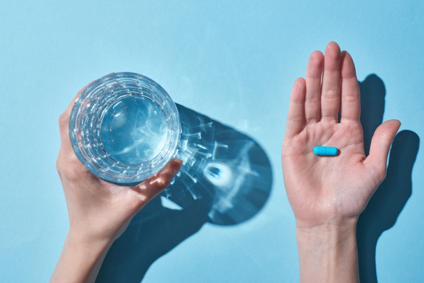 abgeschnittene Ansicht einer Frau mit blauer Pille auf Handfläche und Glas mit Wasser auf blauem Hintergrund - Foto, Bild