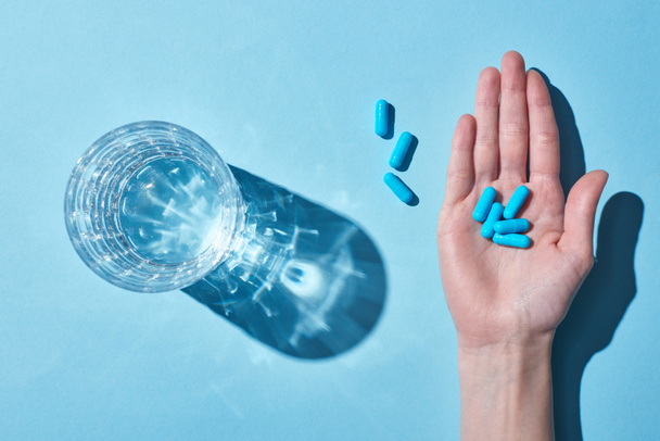 青い背景に水とガラスの近くの手のひらに青い錠剤を保持している女性のトリミングビュー - 写真・画像