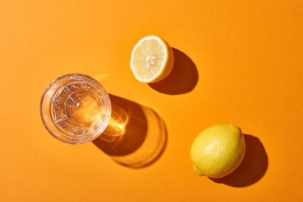 top view of glass with water near yellow lemons on orange background - Фото, изображение
