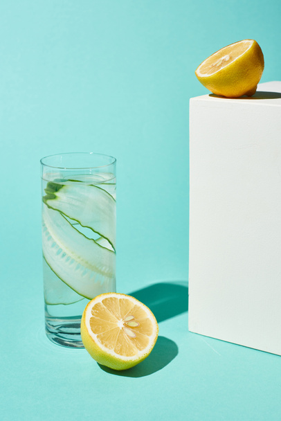 transparent glass with pure water and cucumber slices near lemons on turquoise background - Photo, Image