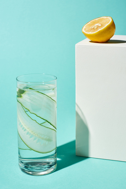 átlátszó üveg tiszta vízzel és uborka szeletekkel közelében citrom fele a türkiz háttér - Fotó, kép