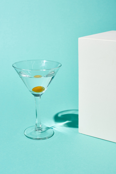 transparent glass with cocktail and olive near white cube on turquoise background - Photo, Image