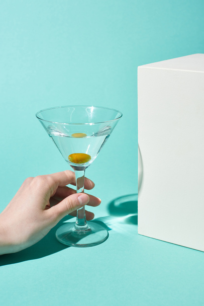 cropped view of woman holding transparent glass with cocktail and olive near white cube on turquoise background - Foto, Imagem