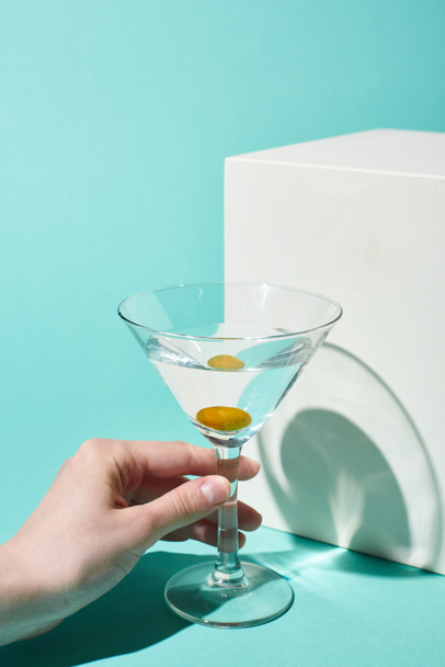 partial view of woman holding transparent glass with cocktail and olive near white cube on turquoise background - Fotografie, Obrázek