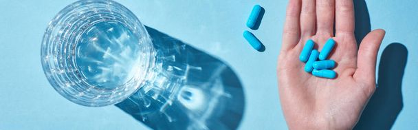 cropped view of woman holding blue pills on palm near glass with water on blue background, panoramic shot - Photo, image