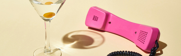 панорамний знімок прозорого скла з коктейлем та оливковою олією біля рожевого старовинного телефону на бежевому фоні
 - Фото, зображення