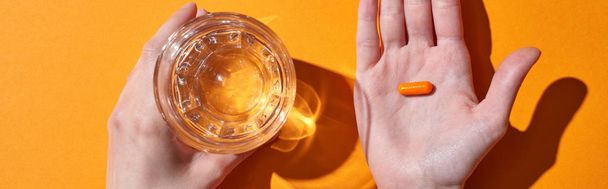 cropped view of woman holding pill on palm near glass with water on orange background, panoramic shot - Foto, Imagem
