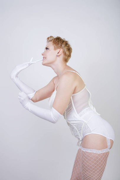 charming caucasian chubby girl with short blonde hair in a cream-colored corsage, stockings and long gloves posing on a white background in the Studio alone - Foto, Imagen
