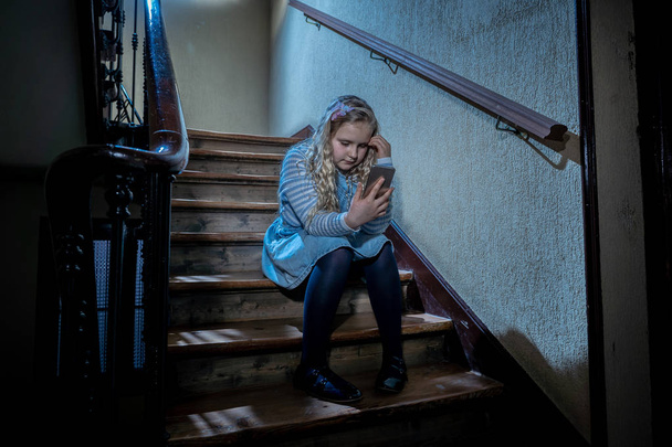 Sad depressed young girl victim of cyberbullying by mobile smart phone sitting on stairs feeling lonely, unhappy, hopeless and abused. Child bullied and harassed by text message by online stalker. - Foto, Imagen