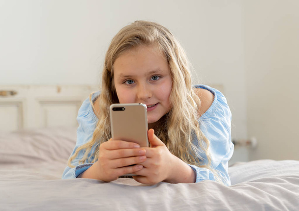 Beautiful blonde happy little girl playing on mobile phone. Cute child chatting on the internet with smartphone social media app lying in bed at home. Technology education and children mobile abuse. - Foto, Bild