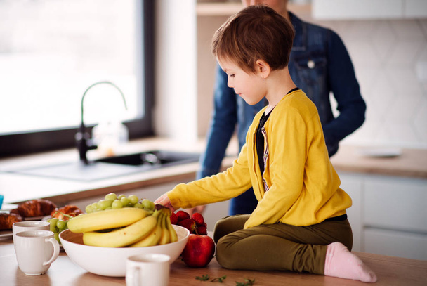 A small girl sitting on the counter in a kitchen, eating fruit. - Foto, imagen