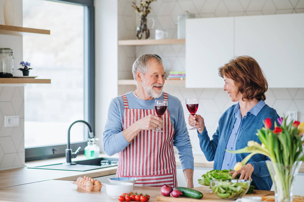 A portrait of senior couple in love indoors at home, clinking glasses when cooking. - Foto, Imagen