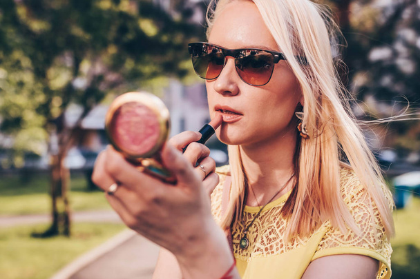 Beautiful blond woman looking in a small mirror and corrects lipstick on the street. - Photo, Image