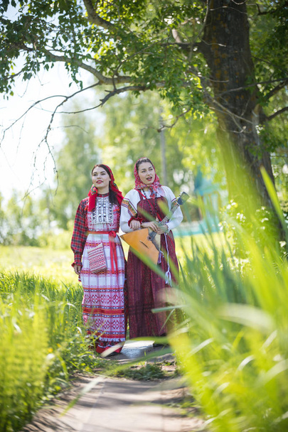 Kaksi nuorta naista perinteisissä venäläisissä vaatteissa kävelemässä polkua pitkin ja puhumassa. Yhdellä heistä oli balalaika.
 - Valokuva, kuva