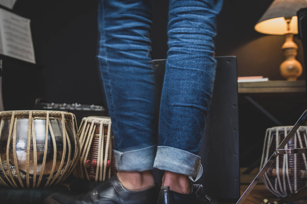 Стильный музыкант в джинсах и туфлях двойного монаха играет на кахоне, перуанском барабане, обычно используемом в испанских танцах фламенко.
.  - Фото, изображение