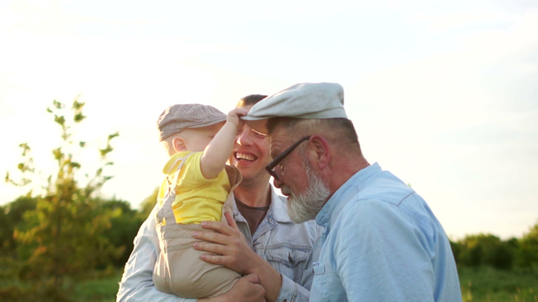 Šťastné miminko na procházce v parku se svou rodinou, otcem a dědečkem. Dítě odstraní čepici od dědečka. Krásný západ slunce, sluneční záře, rodinný koncept - Záběry, video