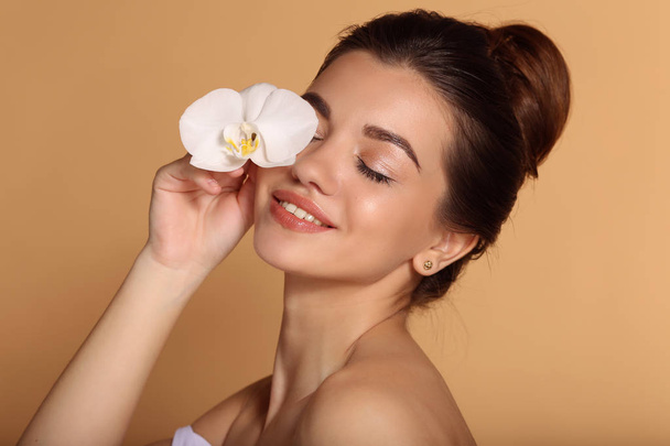 Closeup portrait of young beautiful girl with flawless skin is holding white orchid flower in her hand. Skin care, beauty and cosmetics concept. - Φωτογραφία, εικόνα