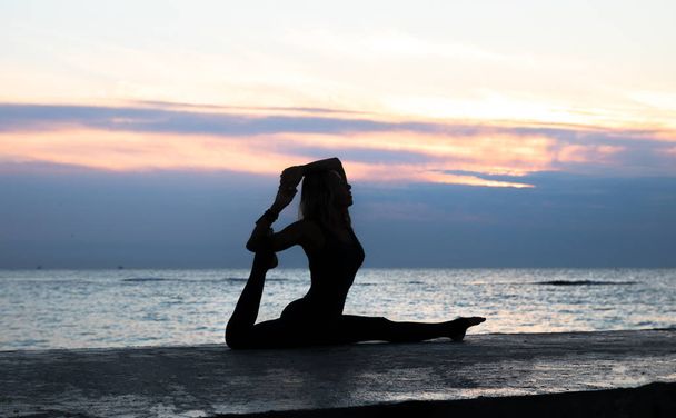 bis zur Unkenntlichkeit senoir Frau mit schönem Körper macht Yoga-Spagat bei Sonnenaufgang über dem Meer, Silhouette von Yoga-Posen - Foto, Bild