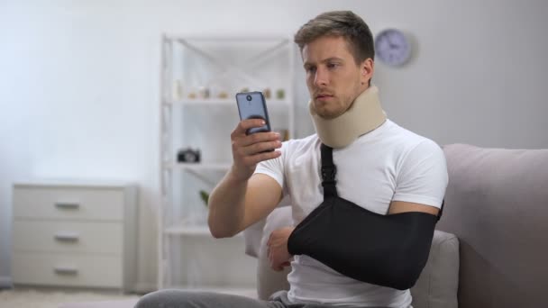 Male in arm sling and foam cervical collar scrolling on smartphone, rehab period - 映像、動画