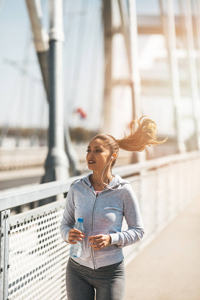Beautiful young and fit woman in good shape running and jogging alone on city bridge street. She wears sporty earbuds and listens to music. - Fotó, kép