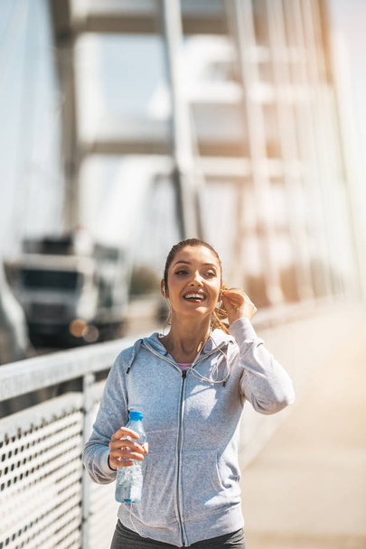 Beautiful young and fit woman in good shape running and jogging alone on city bridge street. She wears sporty earbuds and listens to music. - Foto, Imagem