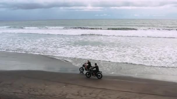 Kaksi komeaa hipsteriä ratsastaa modernilla moottoripyöräkahvilalla mustalla hiekkarannalla lähellä merta. Seikkailumatkakonsepti. Surffauspaikka meren aaltojen kanssa. Avaruuslaukaus. 4 k.
 - Materiaali, video