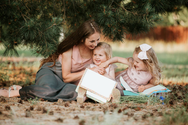 Happy family - mom and two daughters are sitting in a meadow and reading a book. Picnic. happy family, mom and two daughters. Mother's day concept - Φωτογραφία, εικόνα