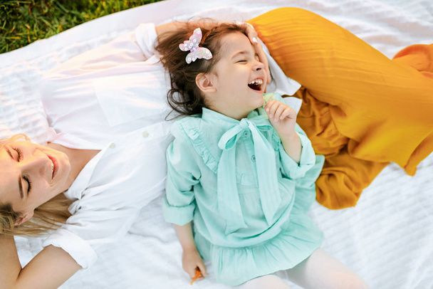 Cute litttle girl playing and having fun with her mother on white blanket at the green grass. Mom and little girl shares love. Mother and daughter smiling and spending time together in the park - Photo, image