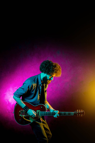 Young hipster man with curly hair with red guitar in neon lights. Rock musician is playing electrical guitar. - Photo, Image