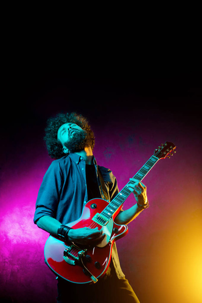 Young hipster man with curly hair with red guitar in neon lights. Rock musician is playing electrical guitar. - 写真・画像