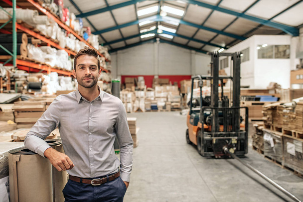 Portrait of a smiling warehouse manager leaning against some stock with piles of carpets on shelves and a forklift in the background - Fotoğraf, Görsel