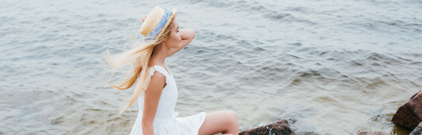 panoramic shot of dreamy blonde girl touching straw hat while sitting in white dress near sea - Photo, Image